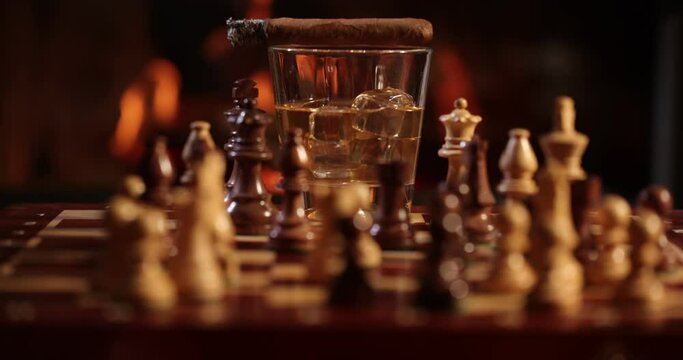Drinks at fireplace on winter evening. Time to enjoy!   High-quality cigars and cognacs, whiskey on a wooden chess table. A flames in the background. Whiskey , chess and steaming cigar on the table in