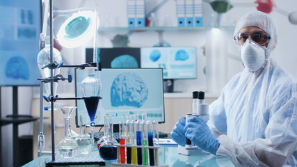 Portrait of chemist researcher with ppe equipment looking into camera while working at microbiology experiment in chemistry hospital laboratory. Scientist doctor analyzing dna sample using microscope