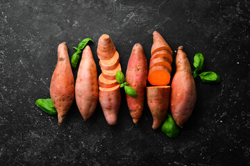 Raw sweet potatoes and basil on a black stone background. Top view. Free space for your text.