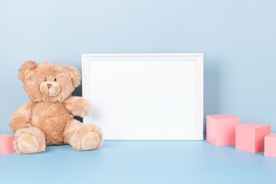 Kids toys collection. White wooden picture frame with blank mock up copy space standing next to teddy bear and pink wooden cubes on light blue background. Front view