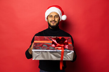 Young indian man in Santa hat holding Christmas gift standing isolated on red background