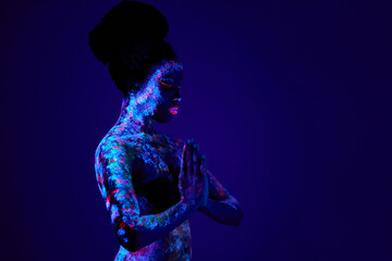 Fototapeta premium young slim black prayer woman with colorful fluorescent prints on skin, beautiful female pray, ask for blessing. prints glows in UV lights. body art, fantasy concept. isolated in studio
