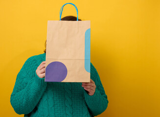 a woman in a sweater holds a paper bag on a yellow background and shows a like gesture with her...