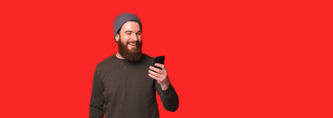 Banner size photo of young cheerful bearded hipster man using smartphone and smiling.