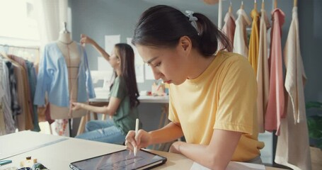 Two professional Asia lady fashion designer with casual use digital tablet create new collection in her shop. Lady tailor study draw sketch on tablet pad and use tape measure in dummy design pattern. - Powered by Adobe