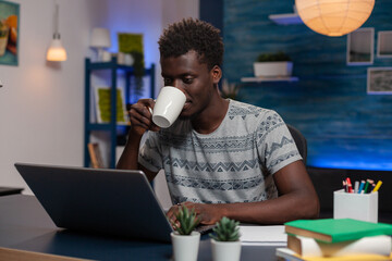 Portrait of african american young entrepreneur sitting at desk typing marketing ideas analyzing financial graph on laptop computer. Employee drinking coffee while working remote from home