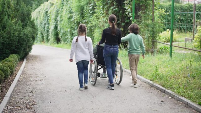 Back view Caucasian children pushing wheelchair with disabled friend outdoors walking in slow motion. Wide shot happy boy and girls taking care of ill paraplegic girl strolling in summer park