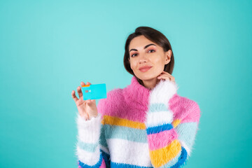 A young woman in a bright multi-colored sweater on a blue background shows a credit card with an...