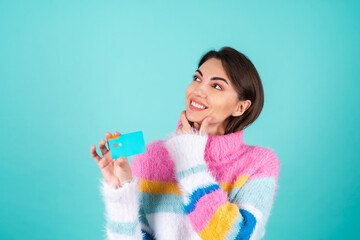A young woman in a bright multicolored sweater on a blue background shows a credit card, looks...