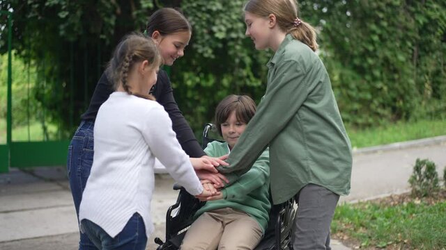 Cute Caucasian disabled boy in wheelchair stacking hands with cheerful girls in slow motion outdoors. Charming friendly children having fun enjoying leisure in summer park. Friendship and disability