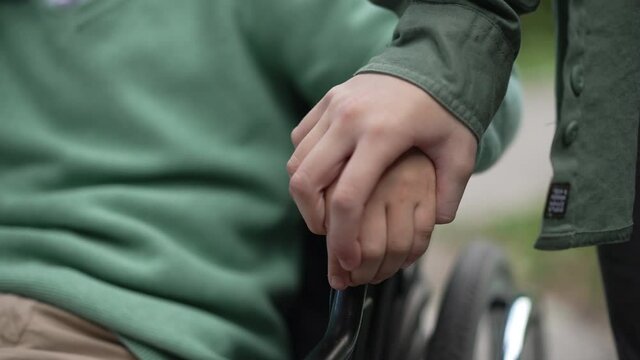 Close-up child hand on wheelchair armrest with unrecognizable girl touching palm of disabled friend in slow motion. Paralyzed Caucasian child outdoors supported by friend. Endorsement concept