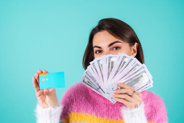 Young woman in a bright multicolored sweater on a blue background shows a credit card and a bundle of fan of dollar money, smiles cheerfully, satisfied
