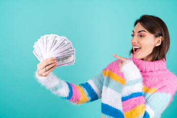 Young woman in a bright multicolored sweater on a blue background holds a bundle of dollars, smiles cheerfully, pointing at money with her finger