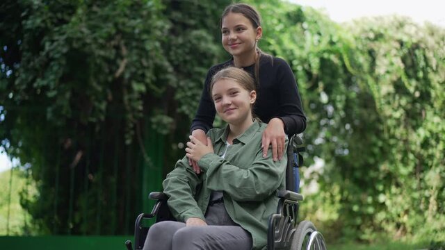 Portrait of happy disabled girl in wheelchair looking at camera smiling resting with friend in summer park. Positive beautiful Caucasian teenager posing in slow motion as friend hugging ill person