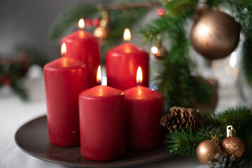 Obraz na płótnie Canvas Christmas decoration with red candles on brown plate, spruce branches, cones, balls and garland on white table on blurry background. Side view. New year mood, festive concept, holiday table, gift card