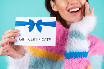 Young woman in a bright multicolored sweater on a blue background holds a gift certificate, smiles...