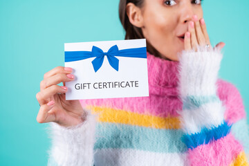 Young woman in a bright multicolored sweater on a blue background holds a gift certificate, smiles...