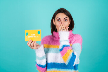 Young woman in a bright multicolored sweater on a blue background holds a gift certificate,...
