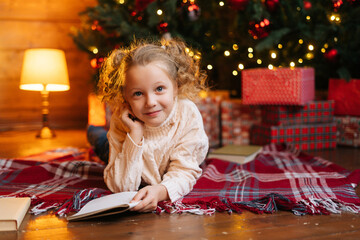 Portrait of happy cute little blonde curly child girl writing letter to Santa Claus lying on floor on background of Christmas tree and gift boxes, looking at camera, in room with festive interior.