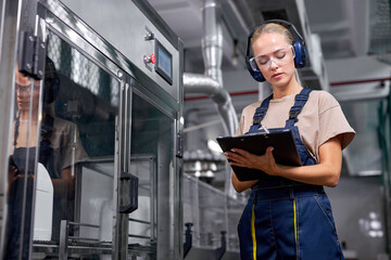 woman worker in headset checking robotic line for bottling and packaging pesticides into bottles and canisters, take notes in folder-tablet , writing with pen on paper. production, manufacturing