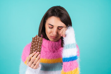 Young woman in a bright multicolored sweater on a blue background suffers, cries, holds a bar of...