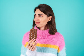 Young woman in a bright multicolored sweater on a blue background suffers, cries, holds a bar of milk chocolate, obesity problems, bulimia