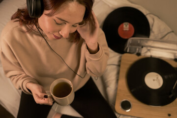 woman sitting on bed listening to music on turntable with headphones and drinking herbal tea and smiling, woman sitting in bed listening to music, woman sitting on bed listening to music on turntable 