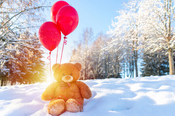 Teddy bear sitting,holding red balloons at cold winter park.Sunset blurred park...