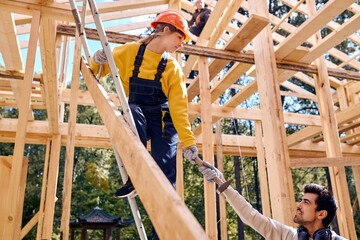 bearded male in uniform helps young caucasian woman constructor standing in height on ladder, in hardhat, bearded responsive male is giving hammer, working as team. friendly and cooperative.