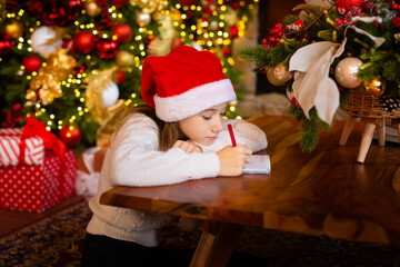 Merry Christmas and Happy Holidays. little child girl in santa claus cap writes letter Santa Claus and dreams of a gift background New Year tree indoors. Greeting card, Christmas mood concept.