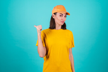 Young woman delivery service worker, courier, in a cap on a blue background smiles broadly and points fingers to the left