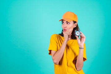 Young woman delivery service worker, courier, in an orange yellow cap on a blue background holds a clock and looks pensively to the side