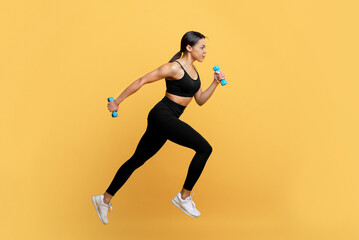 Fototapeta na wymiar Active lifestyle. Fit black lady running with two dumbbells in hands, working out on yellow studio background