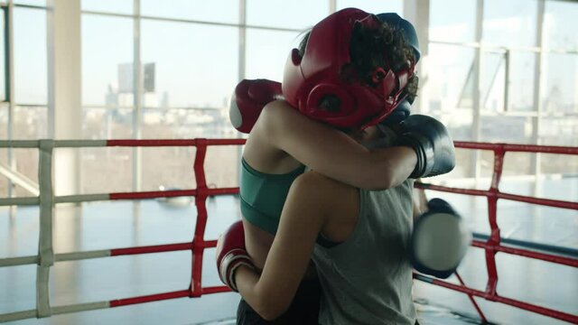 Slow motion of beautiful young women wearing helmets and gloves hugging after match in modern gym smiling enjoying workout together.