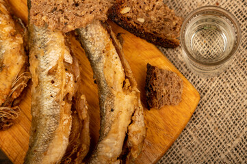 A small fried fish on a wooden board, pieces of bread and a glass of vodka, close-up, selective focus.
