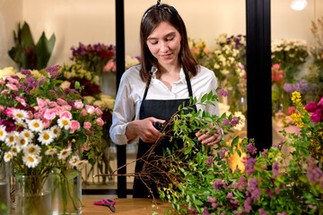 Photo of successful modern florist wearing apron making creating bouquet of blooming colorful different flowers in modern interior floral shop, using scissors. small flower shop business concept