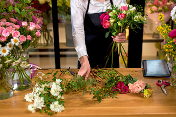 Florist at work: cropped caucasian woman arranging floral decorations, making bouquet for clients. adorable lady in apron is standing behind desk working. floristry, craft and handmade concept