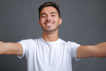 Smiling young handsome man in a white t-shirt looking at camera and making selfie on a gray background