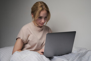 woman browsing social media on laptop in bed, blonde woman shopping online with laptop in bed in the morning