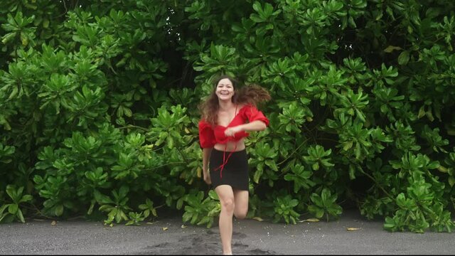 A cheerful young Asian girl with long hair, red blouse and a black mini skirt, jumps against background of huge wall of dawn on beach of Bali. Female traveler smiles with joy and jumps in slow motion.