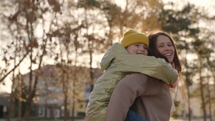 little child is having fun with her mother in city park, happy family life, kid girl on her back mom laughs, childish desires play, daughter love hug mom, laughing cheerful people on street glare sun