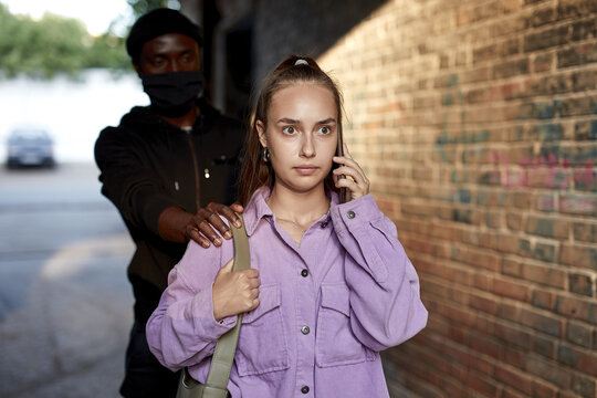 Criminal stalking woman, commiting a crime while victim was walking alone, talking on phone in dark street alley. Caucasian young woman is going to be raped by unrecognizable stranger