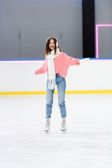 positive young woman in ear muffs and scarf skating with outstretched hands on ice rink.