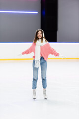 full length of cheerful woman in white ear muffs and scarf skating with outstretched hands on ice rink.