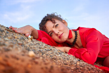 Portrait of a beautiful young woman or girl with curly hair and in a red dress on the sand on sunny day with a blue sky in the background