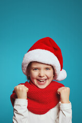 Vertical photo of adorable cute kid wearing christmas hat and red scarf raising fists up and celebrating over blue background 