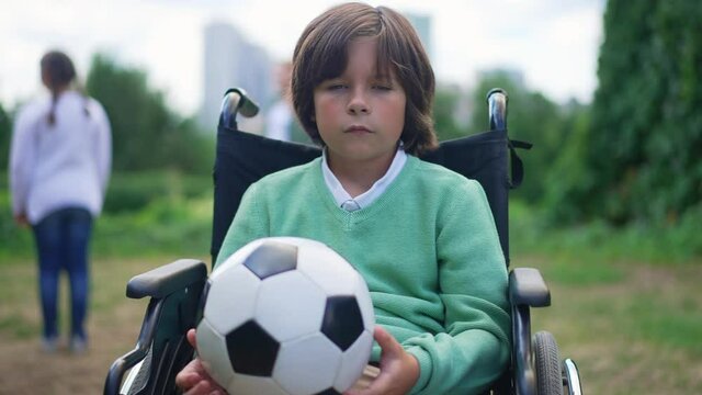 Medium shot sad Caucasian boy with ball in wheelchair thinking and looking at camera in slow motion. Portrait of unhappy upset kid posing in summer park outdoors with children playing at background