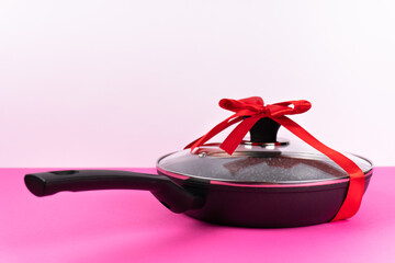New metal frying pan with coating and lid in gift ribbon with bow on pink background. Gift concept...