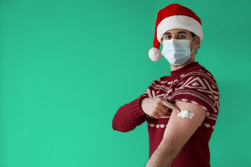 Covid vaccine for Christmas concept. Serious young man with santa hat wearing face mask and red sweater points to bandage in arm over green background
