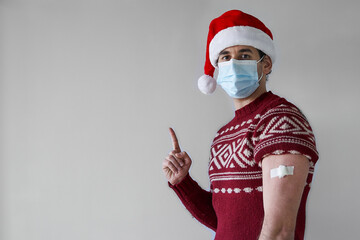 Fototapeta na wymiar Christmas vaccination campaing. Smiling young man with santa hat wearing mask and red sweater with bandage in arm points away over silver grey background